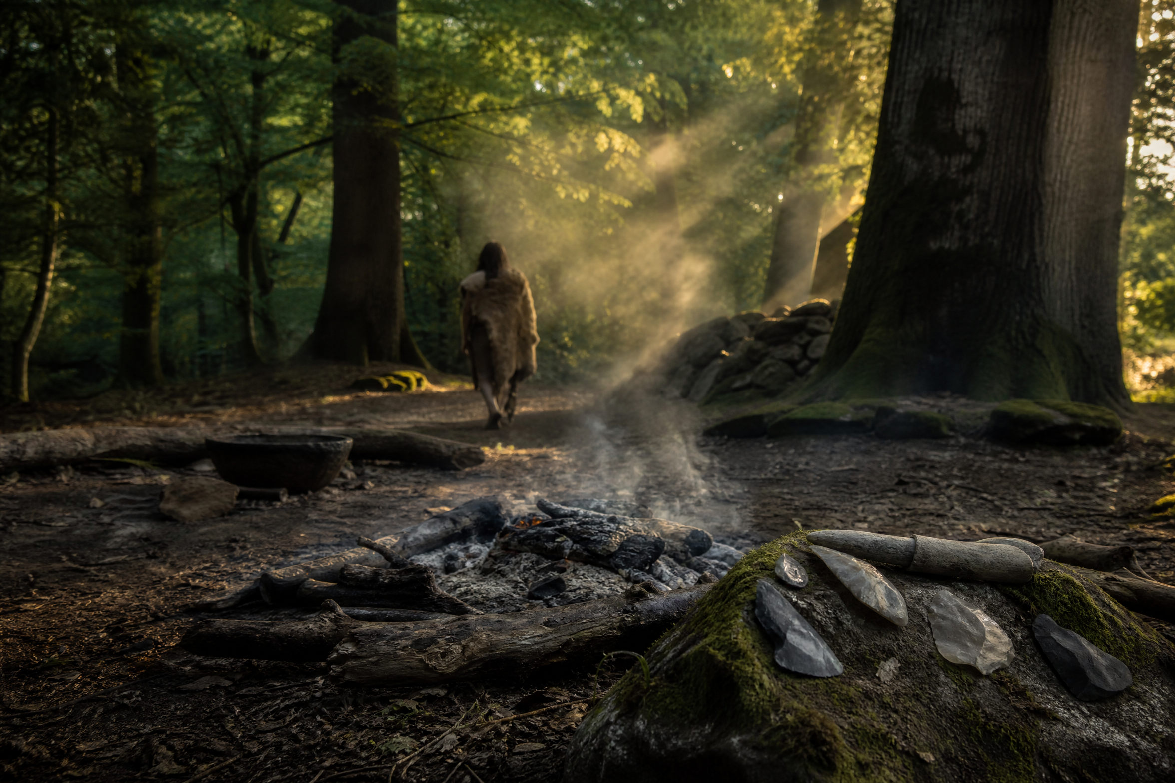 Photorealistic editorial photography of a neolithic-era forest clearing at golden hour, viewed from a low angle: a small smouldering fire ring of charred logs in the foreground, primitive flint hand-tools resting on a mossy stone, a distant